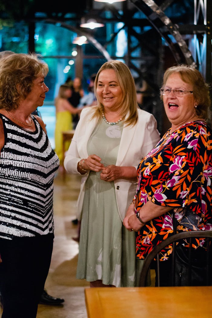 gallery-6 Smiling women engaged in lively conversation at an indoor event, capturing genuine enjoyment.